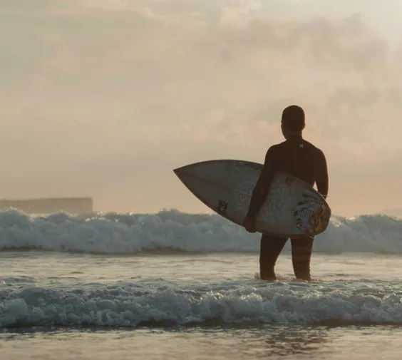 Sober Companion & Coach Taylor Wilson, standing on the beach holding a surfboard